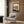 May include: Japanese woodblock print artwork displayed in modern reading nook with cream armchair, geometric pillow, brass floor lamp, and dark round side table with open book. The framed print shows traditional interior scene with wooden lattice window, tatami mats, and mountain landscape view in vintage ukiyo-e style with red, blue, and green colors against warm wood paneled wall.