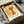 May include: Framed art print featuring an abstract orange cat with yellow eyes and whiskers on aged, textured golden background. The minimalist line drawing shows a stylized feline with pointed ears and curled tail. Print appears to be matted in white with black frame, nestled in shipping box with protective wrapping paper.