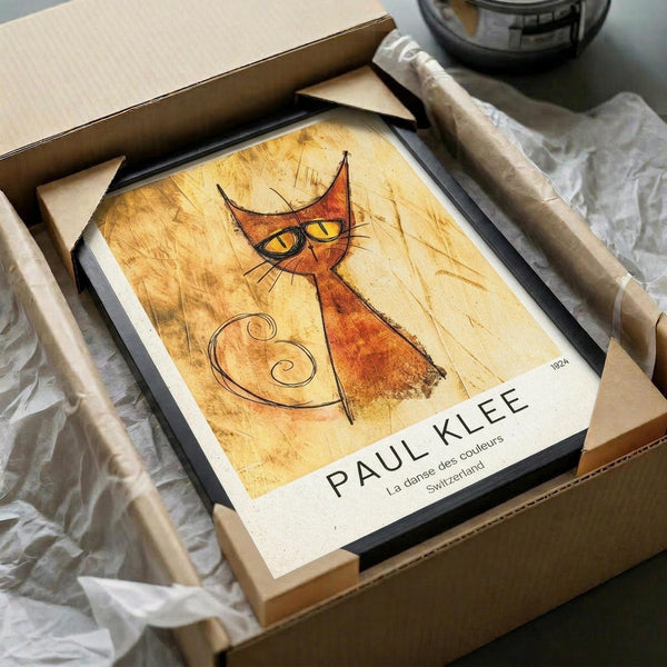 May include: Framed art print featuring an abstract orange cat with yellow eyes and whiskers on aged, textured golden background. The minimalist line drawing shows a stylized feline with pointed ears and curled tail. Print appears to be matted in white with black frame, nestled in shipping box with protective wrapping paper.