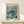 May include: Framed poster featuring a sea turtle swimming among coral in muted green and teal tones, displayed on a light wood console table next to a small wrapped gift with twine bow against a cream wall