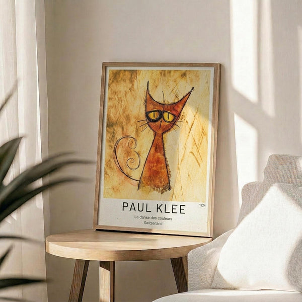May include: Framed poster featuring a stylized orange and red cat with pointed ears, yellow eyes, and curly tail on a golden textured background, displayed on a round wooden side table next to a beige upholstered chair with a white throw pillow, green plant leaves visible in corner, natural sunlight casting shadows on cream colored wall
