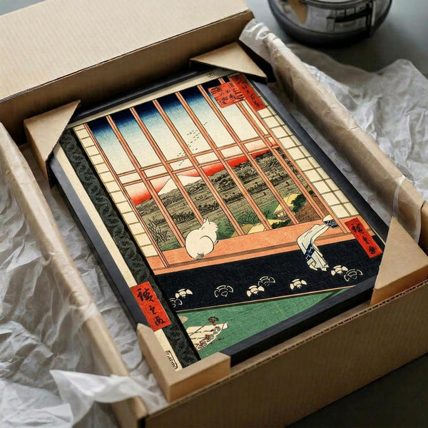 Japanese woodblock print showing a white cat sitting on a wooden veranda overlooking a scenic landscape view through wooden lattice windows. The artwork features traditional ukiyo-e style with vibrant colors including orange sunset sky, green fields, and red text cartouches. The framed print is carefully packaged in a cardboard box with protective wrapping, surrounded by white tissue paper on a neutral surface.