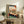 May include: Japanese woodblock print artwork in light wooden frame showing interior scene with sliding doors opening to landscape view with Mount Fuji, white cat on windowsill, displayed on rustic wooden sideboard with books, ceramic vase, and fern plant in modern living room setting with beige chair and neutral walls.