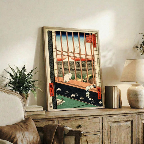 May include: Japanese woodblock print artwork in light wooden frame showing interior scene with sliding doors opening to landscape view with Mount Fuji, white cat on windowsill, displayed on rustic wooden sideboard with books, ceramic vase, and fern plant in modern living room setting with beige chair and neutral walls.