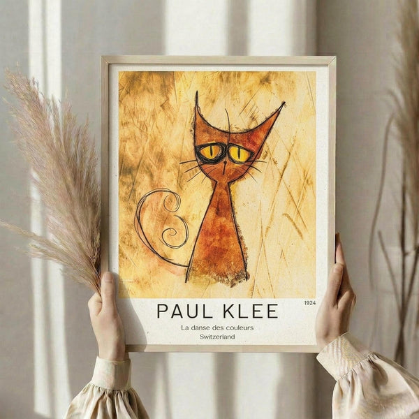 May include: Hands holding a framed art print featuring a stylized orange cat with pointed ears, yellow eyes, and a curly tail against a textured golden background. The print shows "PAUL KLEE" and "La danse des couleurs Switzerland 1924" text below the artwork. Person wears a cream ruffled sleeve top and holds dried wheat stems, standing against white curtains in soft natural lighting.