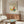 May include: Modern living room with walnut credenza displaying framed Picasso-style poster featuring colorful abstract butterfly with yellow, orange, and teal wings on beige background. White mushroom table lamp, curved white chair, glass-top coffee table with blue and yellow artwork underneath. Warm natural lighting creates shadows on cream walls.
