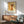 May include: Mid-century modern living room with stylized orange cat poster featuring yellow eyes and curled tail on textured golden background, displayed in thin wooden frame above walnut sideboard with white ceramic lamp, next to sage green upholstered armchair with wooden frame, natural sunlight casting shadows across white walls