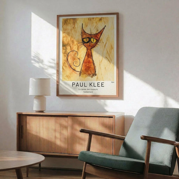 May include: Mid-century modern living room with stylized orange cat poster featuring yellow eyes and curled tail on textured golden background, displayed in thin wooden frame above walnut sideboard with white ceramic lamp, next to sage green upholstered armchair with wooden frame, natural sunlight casting shadows across white walls