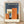 May include: Framed Bauhaus-style architectural poster with geometric orange, cream, blue and brown shapes arranged like abstract buildings on dark blue background. Modern minimalist design in light wood frame, displayed on wooden floor against white wall with natural lighting.