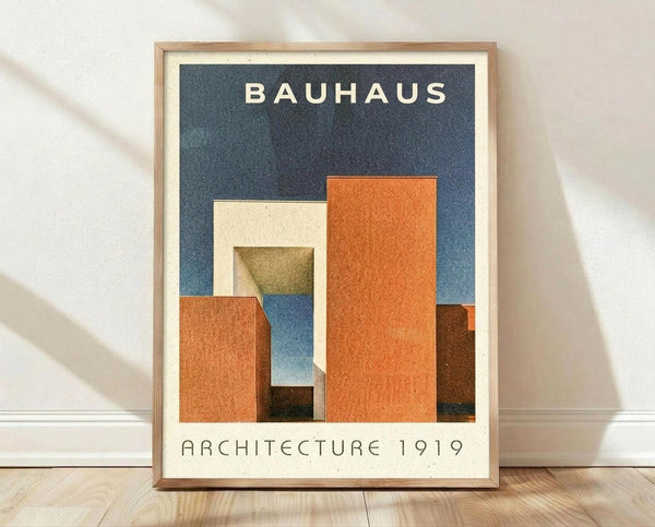 May include: Framed Bauhaus-style architectural poster with geometric orange, cream, blue and brown shapes arranged like abstract buildings on dark blue background. Modern minimalist design in light wood frame, displayed on wooden floor against white wall with natural lighting.