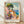 May include: Framed travel poster print featuring a vibrant Mediterranean coastal scene with colorful buildings in pink, orange and yellow, large green tree, stone steps, flowering plants in pots, and blue ocean with sailboat. Painted in impressionistic style with bold brushstrokes. Light wood frame with white matting, leaning against white wall on wooden floor with natural lighting casting shadows.