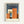 Framed poster with geometric abstract buildings in orange, cream, and brown tones against a dark blue background. The word "BAUHAUS" appears in white letters at the top, with "ARCHITECTURE 1919" at the bottom. The artwork features angular, overlapping architectural forms creating depth and shadow. Displayed in a natural wood frame with white matting against a light gray wall.