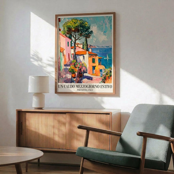 May include: Framed vintage-style travel poster showing colorful Mediterranean coastal village with pink and orange buildings, large green tree, and blue sea. Displayed above wooden credenza with white ceramic lamp in modern living room setting with sage green upholstered armchair and small round side table. Natural sunlight creates shadows on white wall.