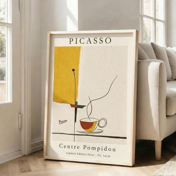 May include: Framed Picasso-style minimalist poster leaning against white wall next to beige sofa in bright living room with large windows. Abstract artwork features yellow geometric shape, simple line-drawn coffee cup with steam, and red accent on cream background. Natural wood frame, museum exhibition text at bottom.