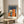 May include: Framed Bauhaus poster with geometric orange, blue, and cream architectural shapes on dark blue background, displayed on round wooden side table next to beige upholstered chair with white pillow, plant leaves visible in corner, natural lighting creating soft shadows on neutral wall