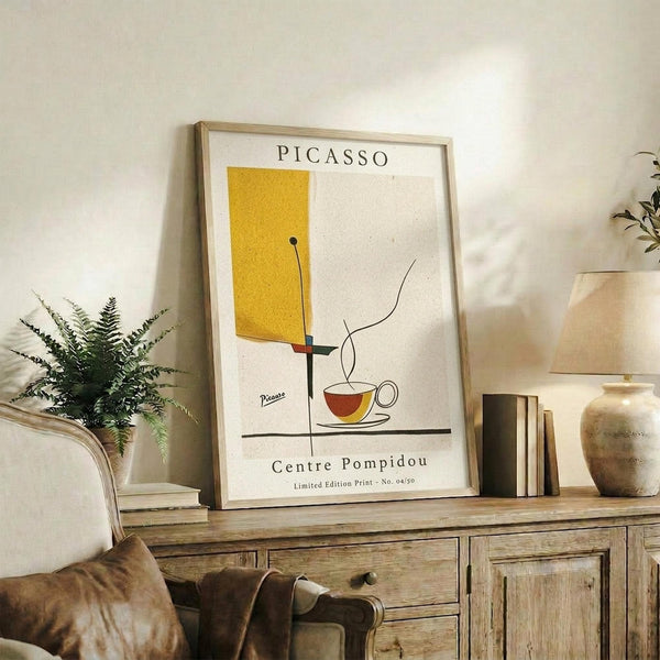 May include: Framed Picasso-style minimalist poster with yellow geometric shape, black lines, and red coffee cup on cream background, displayed on rustic wooden sideboard alongside fern plant, ceramic vases, and books in warm-toned living room setting
