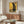 May include: Framed art print displayed above wooden sideboard in modern living room. Artwork features black cat in golden Art Nouveau style with geometric patterns and circular motifs. Room includes white curved chair, glass-top coffee table, and floor lamp. Warm lighting creates reflections on furniture surfaces.