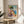 May include: Framed vintage-style poster featuring colorful Mediterranean coastal scene with pastel buildings, large tree, and blue water with sailboat. Print displays in thin wooden frame on round wooden side table next to beige upholstered chair. Soft natural lighting creates window shadows on cream wall. Green plant leaves visible in corner.