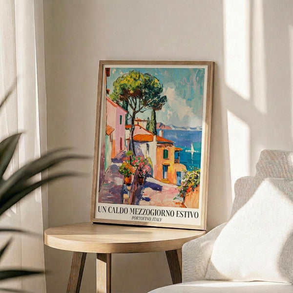 May include: Framed vintage-style poster featuring colorful Mediterranean coastal scene with pastel buildings, large tree, and blue water with sailboat. Print displays in thin wooden frame on round wooden side table next to beige upholstered chair. Soft natural lighting creates window shadows on cream wall. Green plant leaves visible in corner.