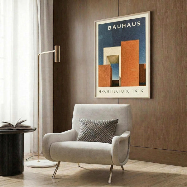 May include: Modern living room with beige armchair featuring geometric patterned pillow, brass floor lamp, and framed Bauhaus poster on wood-paneled wall. Poster displays abstract architectural forms in orange, blue, and cream colors with "BAUHAUS" and "ARCHITECTURE 1919" text on navy background in light wood frame.