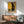 May include: Framed art print featuring a black cat in Gustav Klimt's golden decorative style with ornate circles and patterns on a golden yellow background, displayed above a wooden sideboard with white ceramic lamp in a modern living room setting with teal upholstered chair and natural lighting creating geometric shadows on white wall.