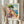May include: Person holding framed vintage-style poster showing colorful Mediterranean coastal scene with pink and orange buildings, blue water, sailboat, and green pine tree. Artwork features painterly brushstrokes in warm sunset tones. Light wood frame against neutral curtained background with dried pampas grass visible.