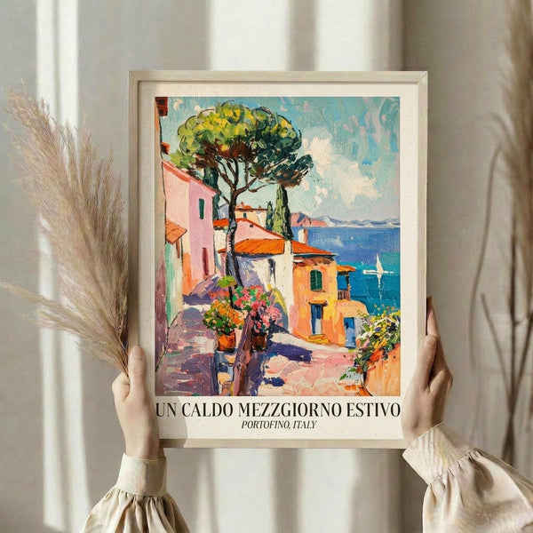 May include: Person holding framed vintage-style poster showing colorful Mediterranean coastal scene with pink and orange buildings, blue water, sailboat, and green pine tree. Artwork features painterly brushstrokes in warm sunset tones. Light wood frame against neutral curtained background with dried pampas grass visible.