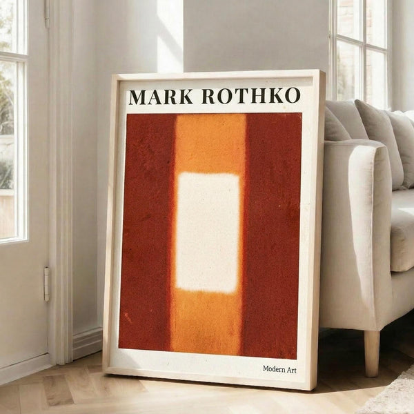 May include: Framed poster print in light wood frame leaning against white wall next to beige sofa. Abstract artwork features deep red background with central vertical rectangle in white and orange tones with soft, blurred edges. Text reads "MARK ROTHKO" at top and "Modern Art" at bottom. Natural lighting creates shadows on hardwood floor in bright, minimalist living room setting.