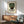 May include: Framed vintage-style poster featuring mosaic mask in green and coral tiles with black silhouette, displayed above wooden sideboard with white ceramic lamp. Mid-century modern living room setting with sage green upholstered chair, round coffee table, and natural sunlight casting geometric shadows on white walls.
