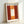 A framed art print featuring a Mark Rothko-style abstract painting with warm tones of deep red, orange, and a glowing white rectangular form in the center. The artwork shows soft, blended color fields characteristic of color field painting. The print is displayed in a light wooden frame and positioned against a white wall, creating gentle shadows that emphasize the frame's depth and texture.