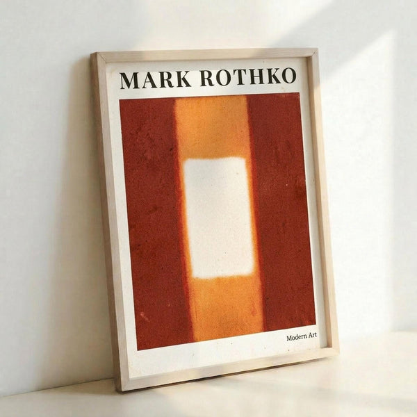 A framed art print featuring a Mark Rothko-style abstract painting with warm tones of deep red, orange, and a glowing white rectangular form in the center. The artwork shows soft, blended color fields characteristic of color field painting. The print is displayed in a light wooden frame and positioned against a white wall, creating gentle shadows that emphasize the frame's depth and texture.