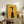 May include: Framed vintage-style poster with yellow background featuring stylized profile of woman in black dress with geometric design. French text at top reads about chic illuminating each detail. Words "prestige" and "chic" in red text. Poster displayed on rustic wooden sideboard alongside books, ceramic vase, and fern plant in neutral-toned living room setting.