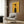 May include: Yellow vintage-style French poster with stylized woman's profile in black hair and striped dress, displaying "prestige" and "chic" text. Shown framed on wood-paneled wall above beige upholstered armchair with geometric pillow, beside brass floor lamp and dark round side table with open book.
