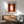 May include: Modern living room with sage green upholstered armchair beside wood sideboard. Framed abstract art print on white wall features warm rust red, orange, and cream rectangular color blocks in minimalist style. White ceramic table lamp and round coffee table complete the mid-century modern interior design scene with natural sunlight streaming across walls.