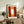 May include: Abstract art poster with orange and red color blocks and white rectangular center, displayed in light wooden frame on rustic wooden sideboard. Room setting shows beige armchair with brown throw pillows, potted fern plant, ceramic vase, and stacked books against cream wall with natural lighting.