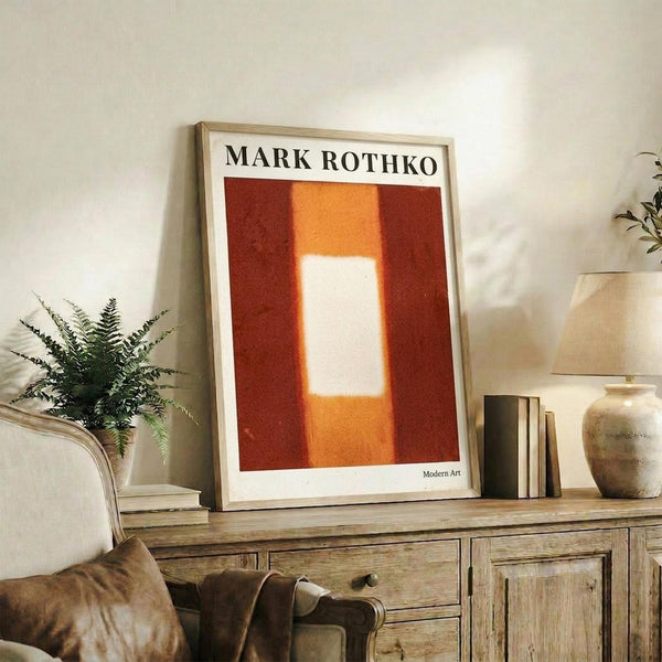 May include: Abstract art poster with orange and red color blocks and white rectangular center, displayed in light wooden frame on rustic wooden sideboard. Room setting shows beige armchair with brown throw pillows, potted fern plant, ceramic vase, and stacked books against cream wall with natural lighting.