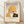 May include: Framed vintage-style art print leaning against white wall on light wood floor. Shows woman in golden yellow headwrap smelling deep red rose, rendered in warm earth tones with Art Deco influence. Features French text "Le Parfum De La Rose" below illustration. Natural wood frame with cream matting, soft window light casting shadows across white textured wall.