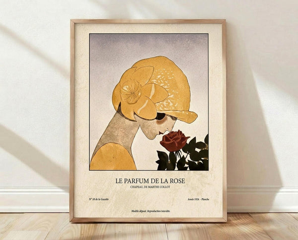 May include: Framed vintage-style art print leaning against white wall on light wood floor. Shows woman in golden yellow headwrap smelling deep red rose, rendered in warm earth tones with Art Deco influence. Features French text "Le Parfum De La Rose" below illustration. Natural wood frame with cream matting, soft window light casting shadows across white textured wall.
