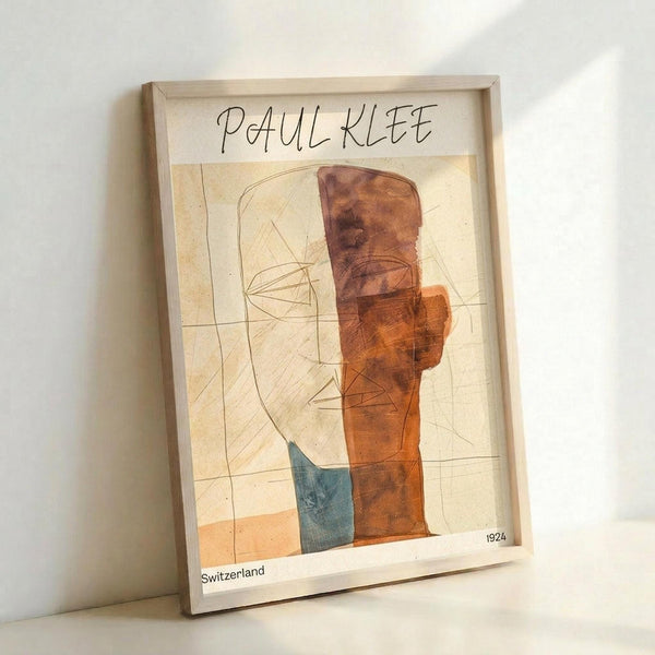 May include: A framed abstract art print leaning against a white wall on a white surface. The artwork features geometric shapes in warm orange and brown tones with a small blue accent, combined with delicate line drawings on an aged cream background. The piece is titled "PAUL KLEE" at the top with "Switzerland" and "1924" at the bottom. The print is displayed in a thin light wood frame.