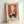 May include: Framed vintage-style poster print leaning against white wall. Orange and cream artwork shows cats around a table by a window with blooming tree branches. Features black silhouettes of cats, green vase, and French text at bottom. Light wooden frame with warm lighting casting soft shadows.