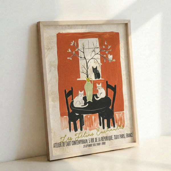May include: Framed vintage-style poster print leaning against white wall. Orange and cream artwork shows cats around a table by a window with blooming tree branches. Features black silhouettes of cats, green vase, and French text at bottom. Light wooden frame with warm lighting casting soft shadows.