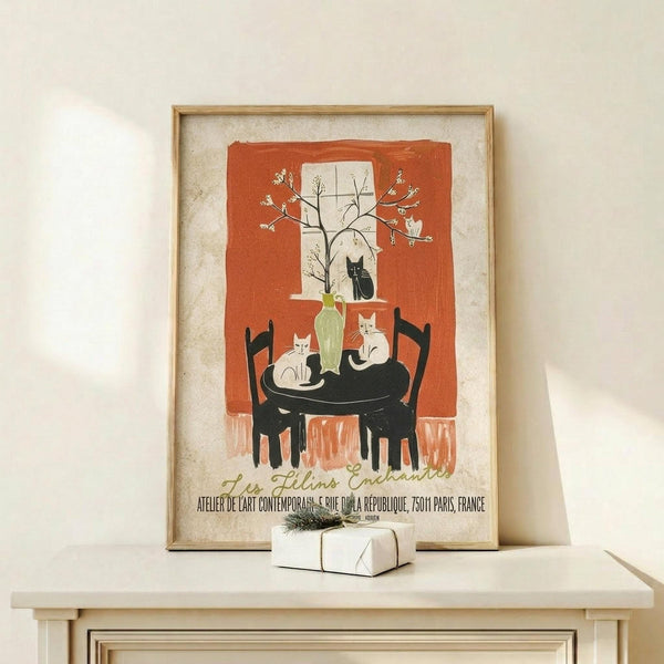 May include: Framed vintage-style poster featuring illustrated cats around a black table with chairs, set against an orange and cream background with a window and flowering branch. The artwork shows a cozy interior scene with a green vase centerpiece. Frame appears in light wood, displayed on a white shelf next to a small wrapped gift with greenery.