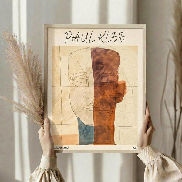 May include: Person in beige clothing holding framed Paul Klee abstract art print featuring geometric shapes in warm orange, rust, and blue tones with delicate line drawings on cream background. Light wooden frame, pampas grass decoration, soft natural lighting against neutral curtained backdrop.