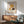 May include: Art Deco style poster showing woman in golden cloche hat smelling dark red rose, displayed in thin wooden frame above mid-century modern wooden credenza with white ceramic lamp, beside sage green upholstered armchair with wooden arms in sunlit room with white walls
