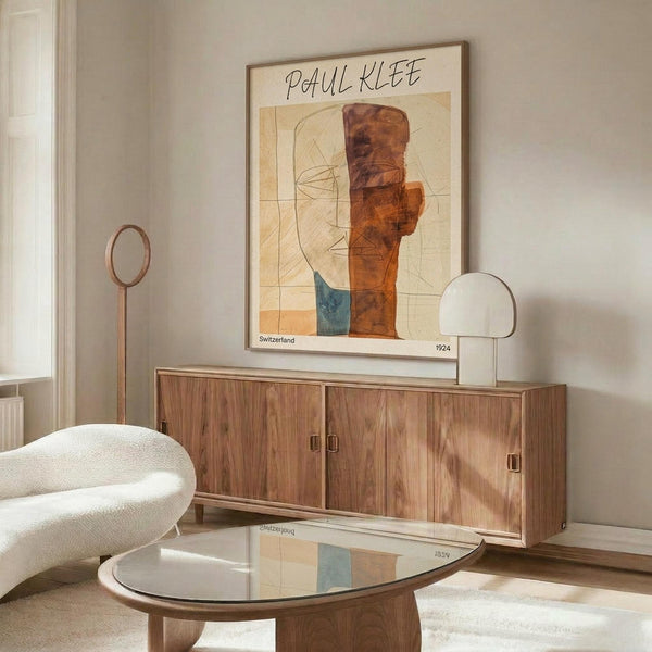 May include: Paul Klee abstract art poster in wooden frame hanging above mid-century walnut credenza in modern living room. Artwork features geometric shapes in warm orange, brown and blue tones with linear elements on cream background. Room styled with white curved chair, glass coffee table, and brass floor and table lamps creating soft lighting on neutral walls.