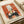 May include: Vintage-style art print displayed on cream paper showing orange and black illustration of cats around a dark wooden table with green vase and bare tree branch against window, featuring French exhibition text at bottom, photographed on white surface with rolled paper in background