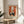 May include: Framed vintage-style poster featuring cats around a black table with green vase and flowering branches against orange background, displayed on light wood round side table next to beige upholstered chair with plant shadows on white wall