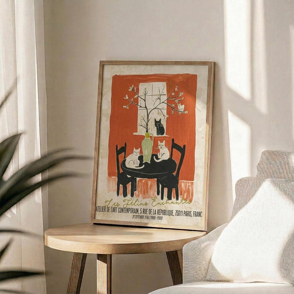 May include: Framed vintage-style poster featuring cats around a black table with green vase and flowering branches against orange background, displayed on light wood round side table next to beige upholstered chair with plant shadows on white wall