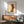 May include: Abstract modernist poster print featuring geometric forms in warm orange, brown, and teal tones with linear face sketches, displayed in wooden frame above mid-century credenza with ceramic table lamp. Styled with sage green upholstered armchair and round coffee table in minimalist living room with white walls and natural sunlight casting shadows.