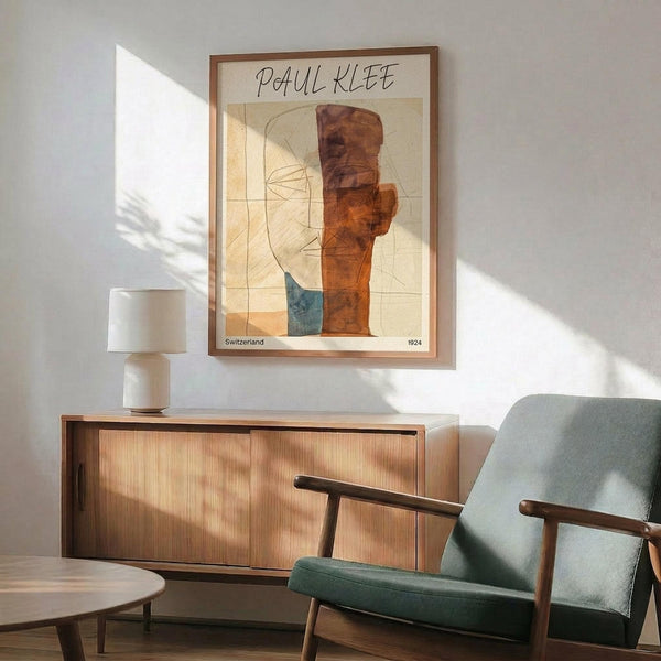 May include: Abstract modernist poster print featuring geometric forms in warm orange, brown, and teal tones with linear face sketches, displayed in wooden frame above mid-century credenza with ceramic table lamp. Styled with sage green upholstered armchair and round coffee table in minimalist living room with white walls and natural sunlight casting shadows.